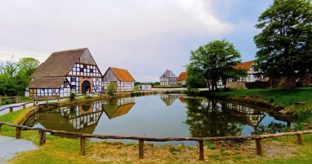 Das Bild zeigt eine malerische ländliche Szene im Freilichtmuseum Detmold mit traditionellen Fachwerkhäusern, die sich auf der Oberfläche eines ruhigen Teichs spiegeln. Ein Holzzaun verläuft im Vordergrund, und üppiges Grün umgibt die Gegend, was die friedliche Atmosphäre der Umgebung verstärkt. Dieses Bild ist interessant, da es eine idyllische Landschaft auf dem Land einfängt, die für Themen wie ländliche Architektur, Geschichte oder Reiseziele relevant sein könnte.
