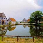 Das Bild zeigt eine malerische ländliche Szene im Freilichtmuseum Detmold mit traditionellen Fachwerkhäusern, die sich auf der Oberfläche eines ruhigen Teichs spiegeln. Ein Holzzaun verläuft im Vordergrund, und üppiges Grün umgibt die Gegend, was die friedliche Atmosphäre der Umgebung verstärkt. Dieses Bild ist interessant, da es eine idyllische Landschaft auf dem Land einfängt, die für Themen wie ländliche Architektur, Geschichte oder Reiseziele relevant sein könnte.