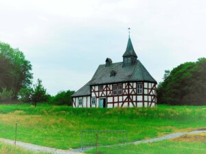 Zu sehen ist eine Kapelle auf einer Wiese die sich auf einer Anhöhe befindet. Die kleine Kapelle ist ein Fachwerkbau mit hellblauen Fenstern und einer Kirchsturmspitze auf der sich ein Wetterhahn befindet.