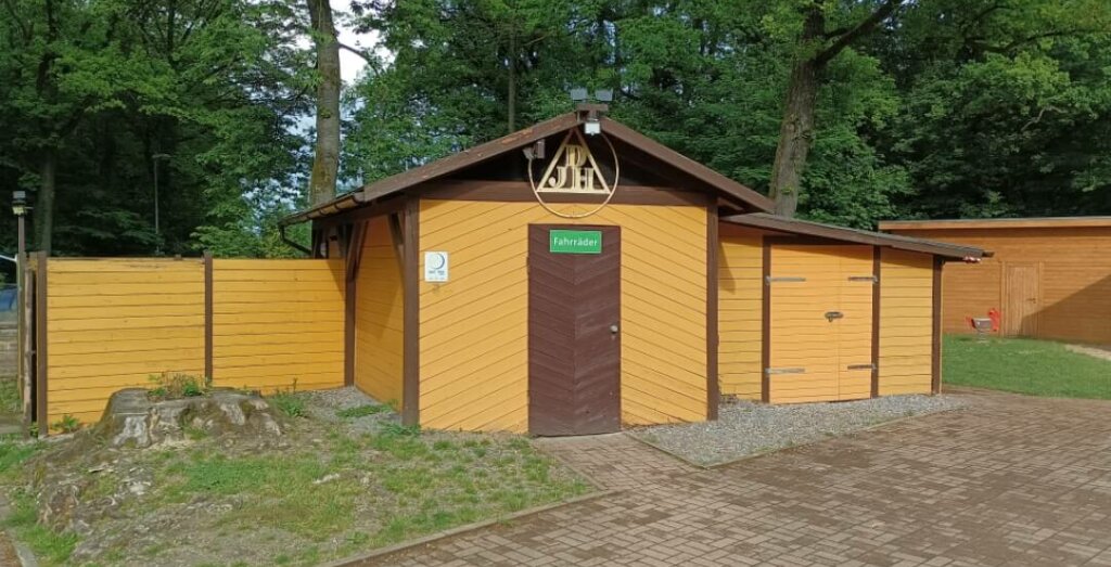 Wir blicken auf das hellbraune Holzfahrradhaus. Über der Eingangstüre des Fahrradschuppens befindet sich ein rundes Schild mit der Aufschrift " DJH".