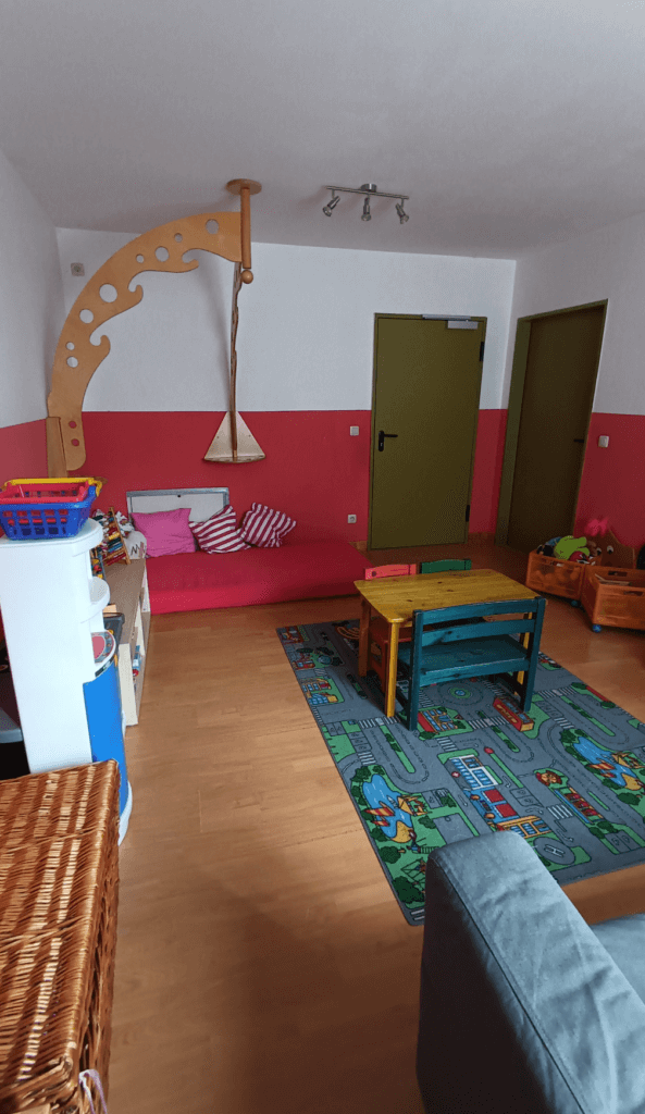 Bild zeigt einen gemütlichen Spiel- und Aufenthaltsraum für Kinder. Der Raum hat eine zweifarbige Wandgestaltung mit Weiß im oberen Bereich und einem rötlichen Farbton im unteren Bereich. Auf der linken Seite befindet sich eine niedrige Spielecke mit einer roten Matratze und mehreren Kissen. Darüber ist eine Holzschaukel mit einem dekorativen, bogenförmigen Aufhängemechanismus angebracht. In der Mitte des Raumes liegt ein farbenfroher Spielteppich mit Straßenmotiven, auf dem sich ein kleiner Holztisch mit bunten Stühlen befindet. An den Wänden sind grüne Türen zu sehen, die den Raum abschließen. Rechts gibt es weitere Spielzeugkisten mit verschiedenen Plüschtieren und anderen Spielmaterialien. Der Raum wirkt gut organisiert und einladend, mit kindgerechten Möbeln und einer freundlichen Farbgestaltung. Bild zeigt einen gemütlichen Spiel- und Aufenthaltsraum für Kinder. Der Raum hat eine zweifarbige Wandgestaltung mit Weiß im oberen Bereich und einem rötlichen Farbton im unteren Bereich. Auf der linken Seite befindet sich eine niedrige Spielecke mit einer roten Matratze und mehreren Kissen. Darüber ist eine Holzschaukel mit einem dekorativen, bogenförmigen Aufhängemechanismus angebracht. In der Mitte des Raumes liegt ein farbenfroher Spielteppich mit Straßenmotiven, auf dem sich ein kleiner Holztisch mit bunten Stühlen befindet. An den Wänden sind grüne Türen zu sehen, die den Raum abschließen. Rechts gibt es weitere Spielzeugkisten mit verschiedenen Plüschtieren und anderen Spielmaterialien. Der Raum wirkt gut organisiert und einladend, mit kindgerechten Möbeln und einer freundlichen Farbgestaltung.