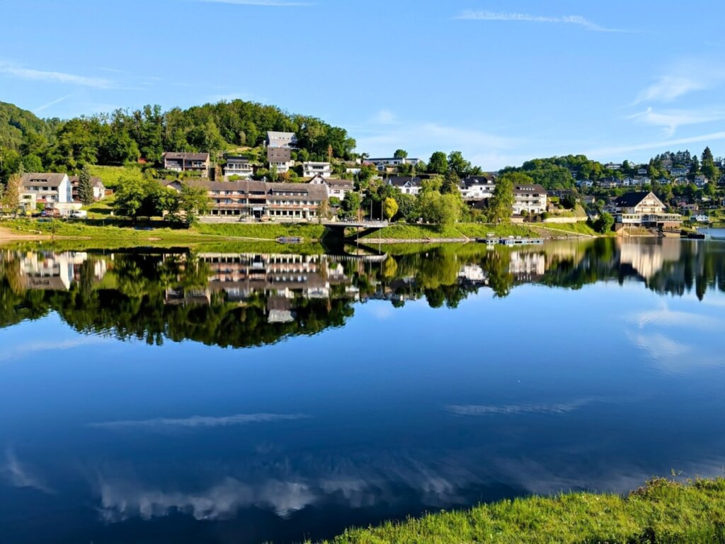 Rurberg am frühen Morgen mit Spiegelung der Häuser im Rursee. Rurberg liegt direkt am Rursee im Nationalpark Eifel und ist ein beliebter Ausgangspunkt für Wanderungen und Naturerlebnisse.