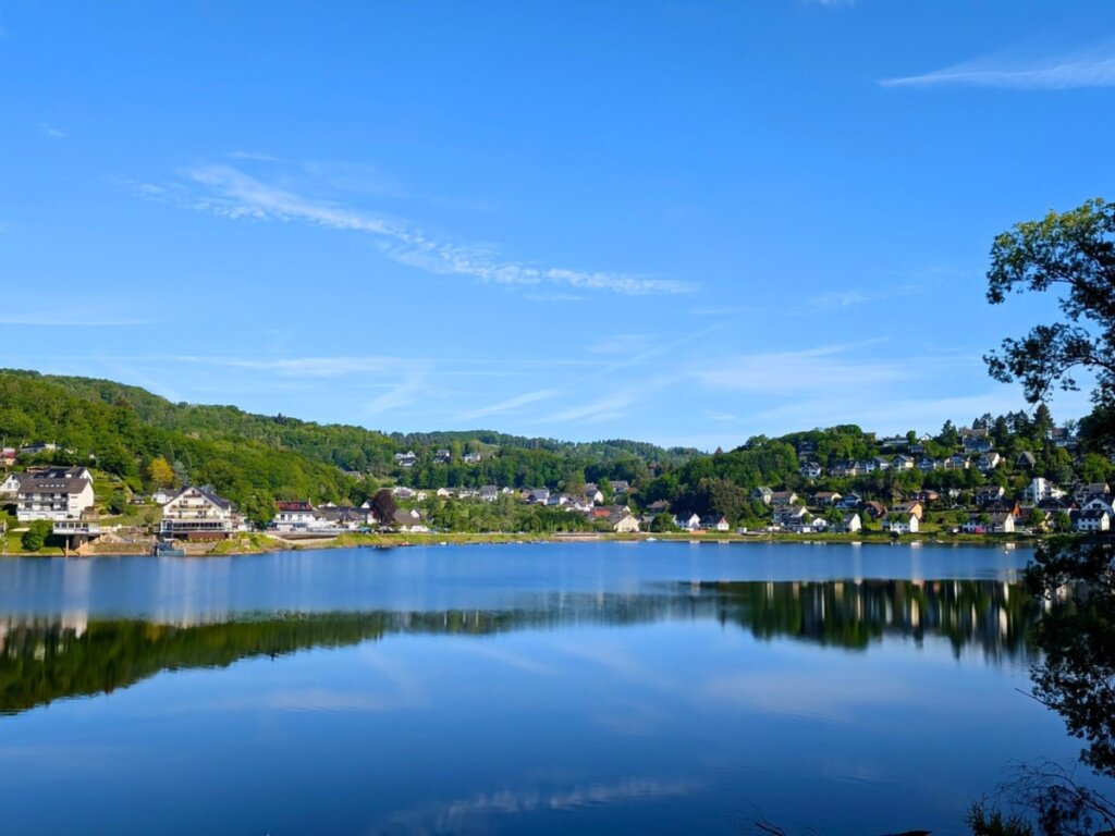 Rurberg am frühen Morgen mit Spiegelung der Häuser im Rursee. Rurberg liegt direkt am Rursee im Nationalpark Eifel und ist ein beliebter Ausgangspunkt für Wanderungen und Naturerlebnisse.