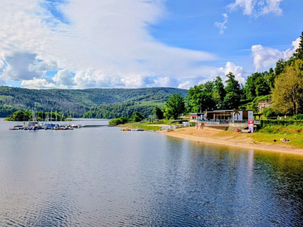 Badestrand Eschauel am Rursee mit Uferbereich, Wasser und bewaldeten Hängen im Hintergrund.